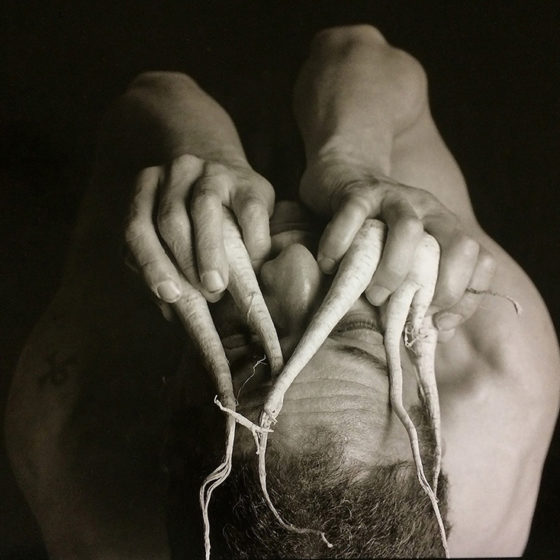 Black and white narritive photograph of man holding roots like fingers over face